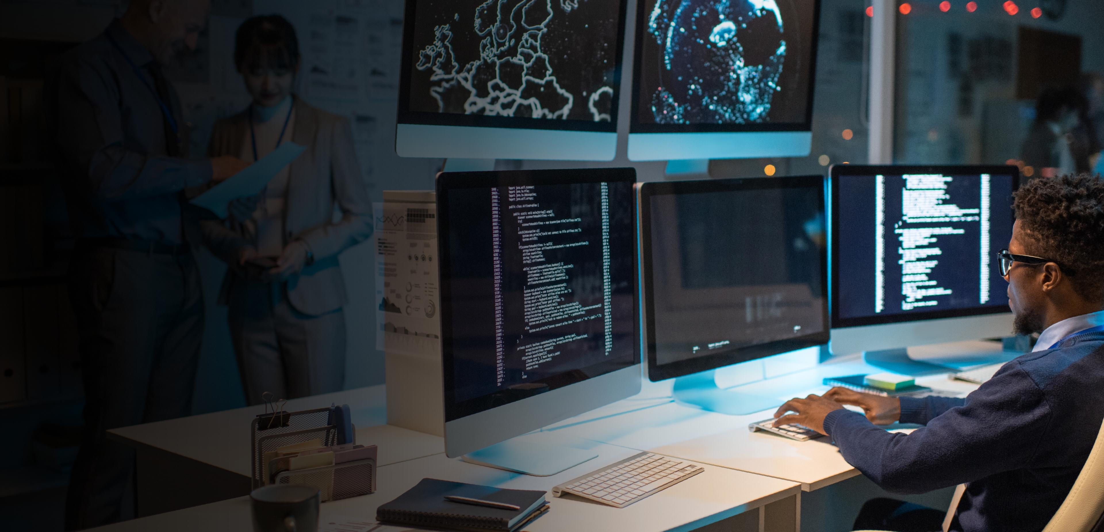 A man seated at a desk, focused on three computer screens displaying various tasks and information.