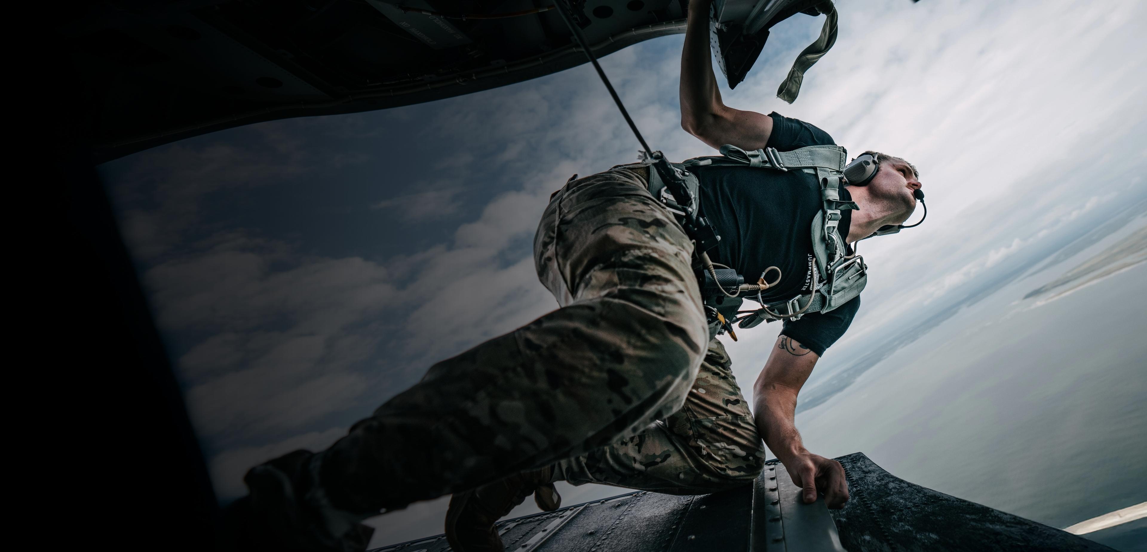 A man in military gear climbs out of a plane, preparing for a mission in a dynamic outdoor setting.