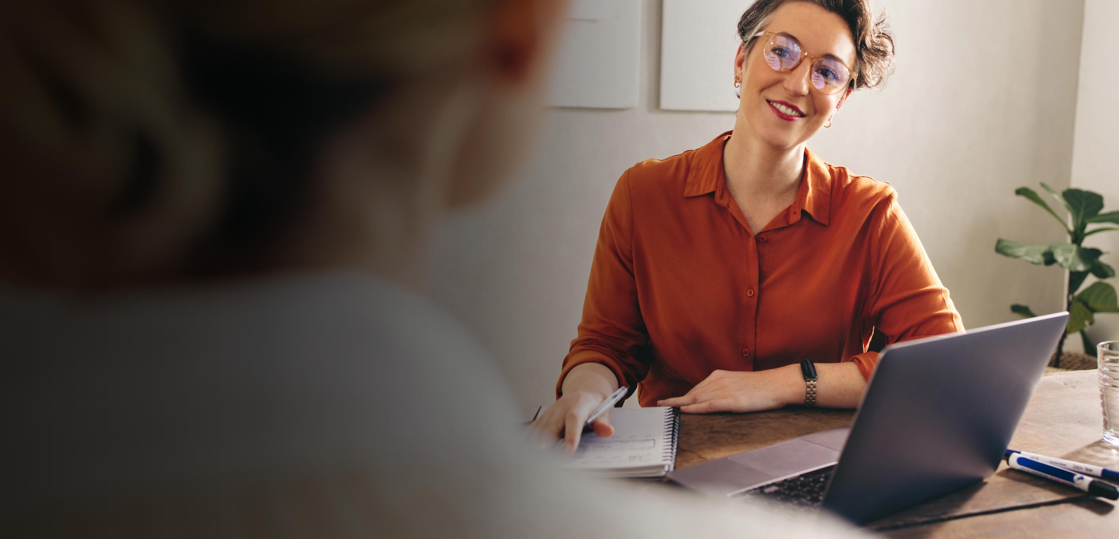 A woman seated at a desk, focused on her laptop, with a notepad and pen beside her.