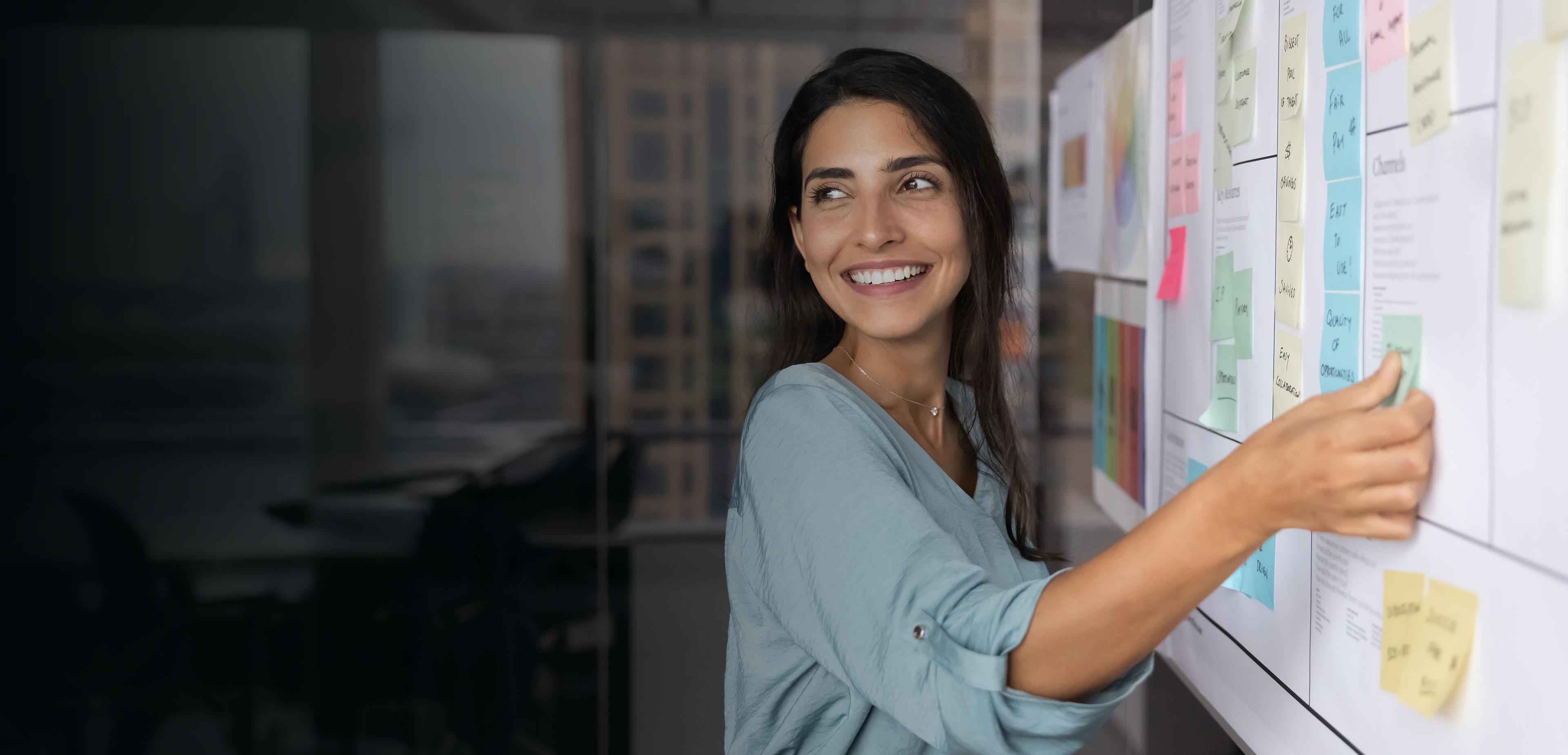 A woman points to a whiteboard covered in colorful sticky notes during a brainstorming session.