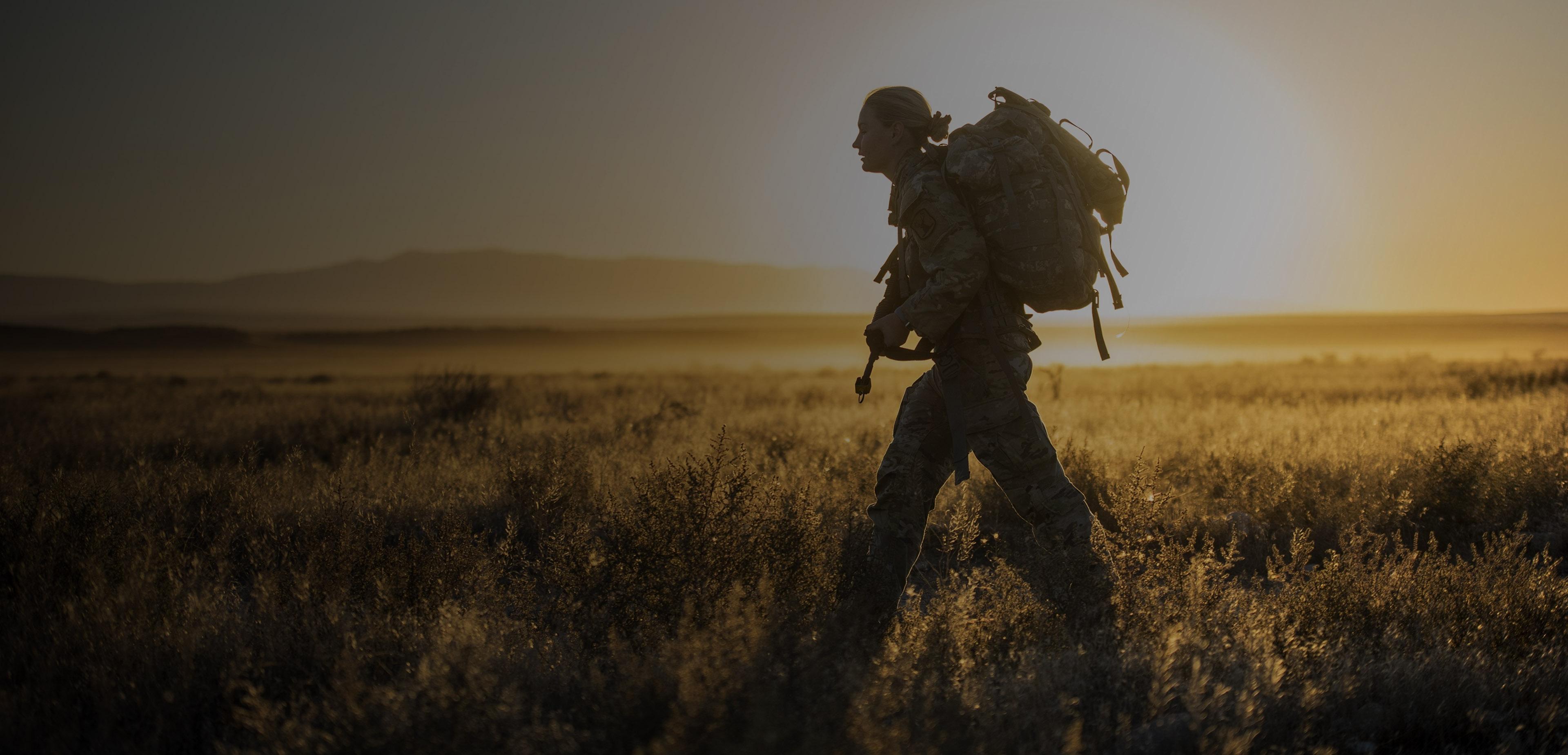 A woman man with a backpack walks through a field during sunset, with warm colors illuminating the landscape around him.