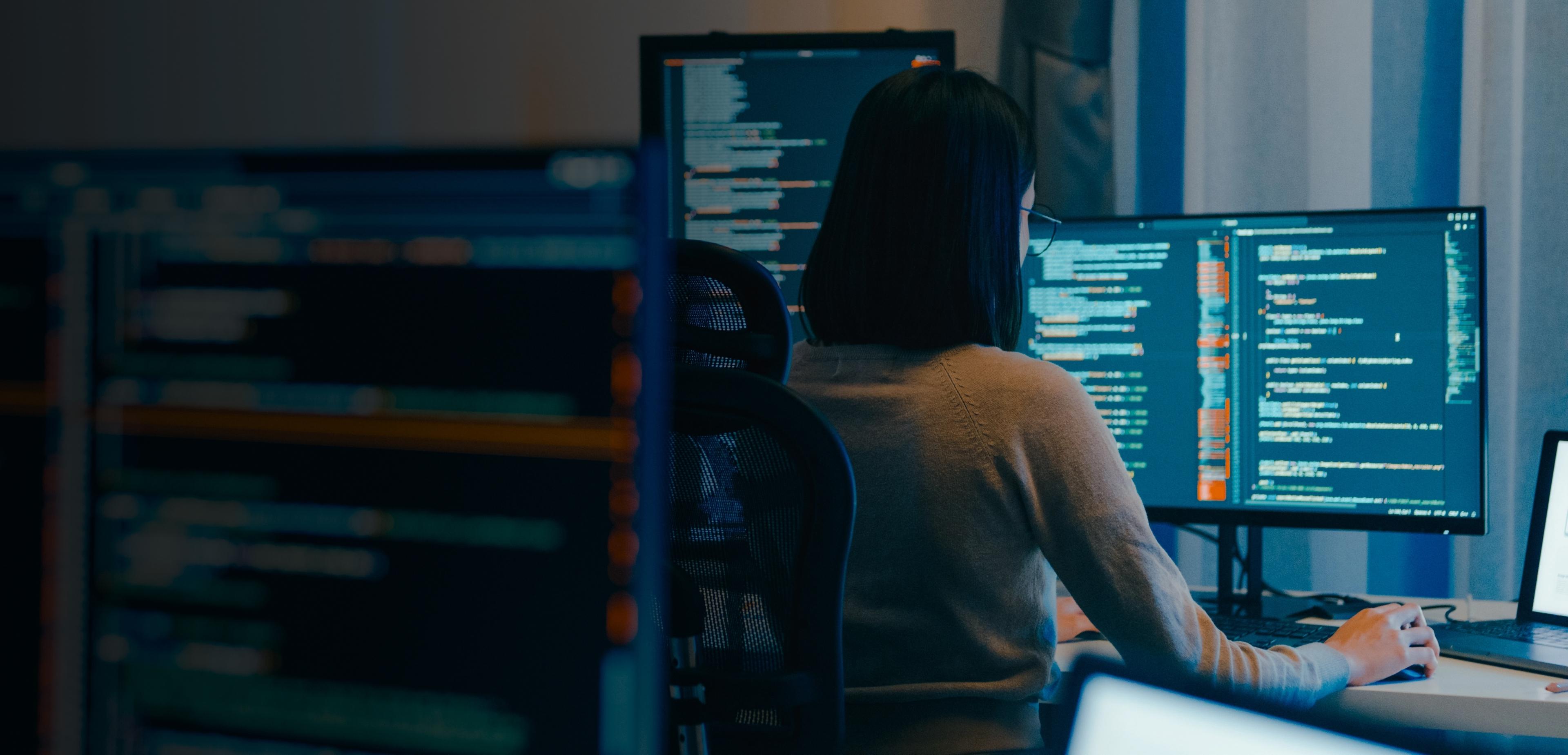 A woman seated at a desk, working on two computers positioned in front of her.