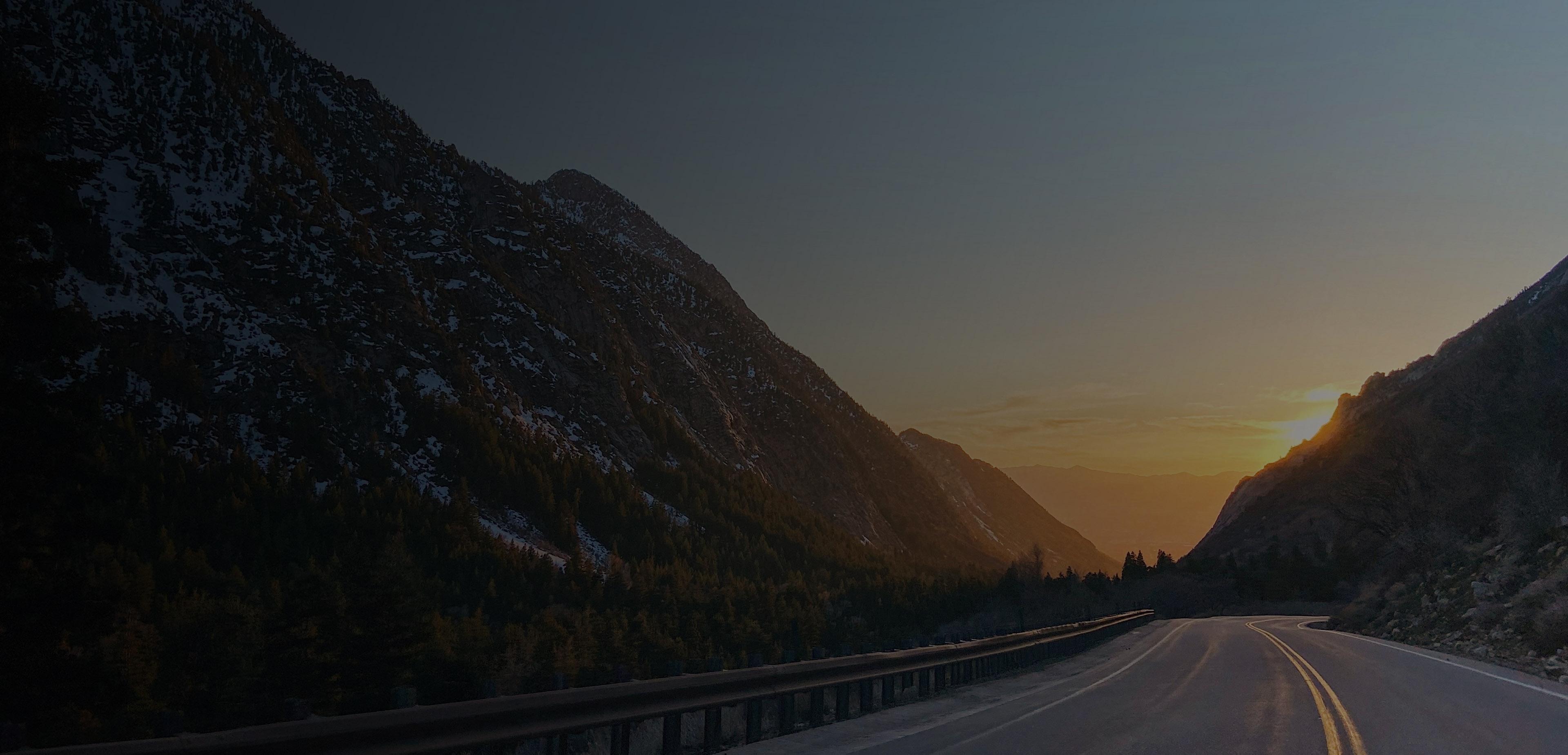 A scenic highway stretches into the distance, framed by majestic mountains under a vibrant sunset sky.