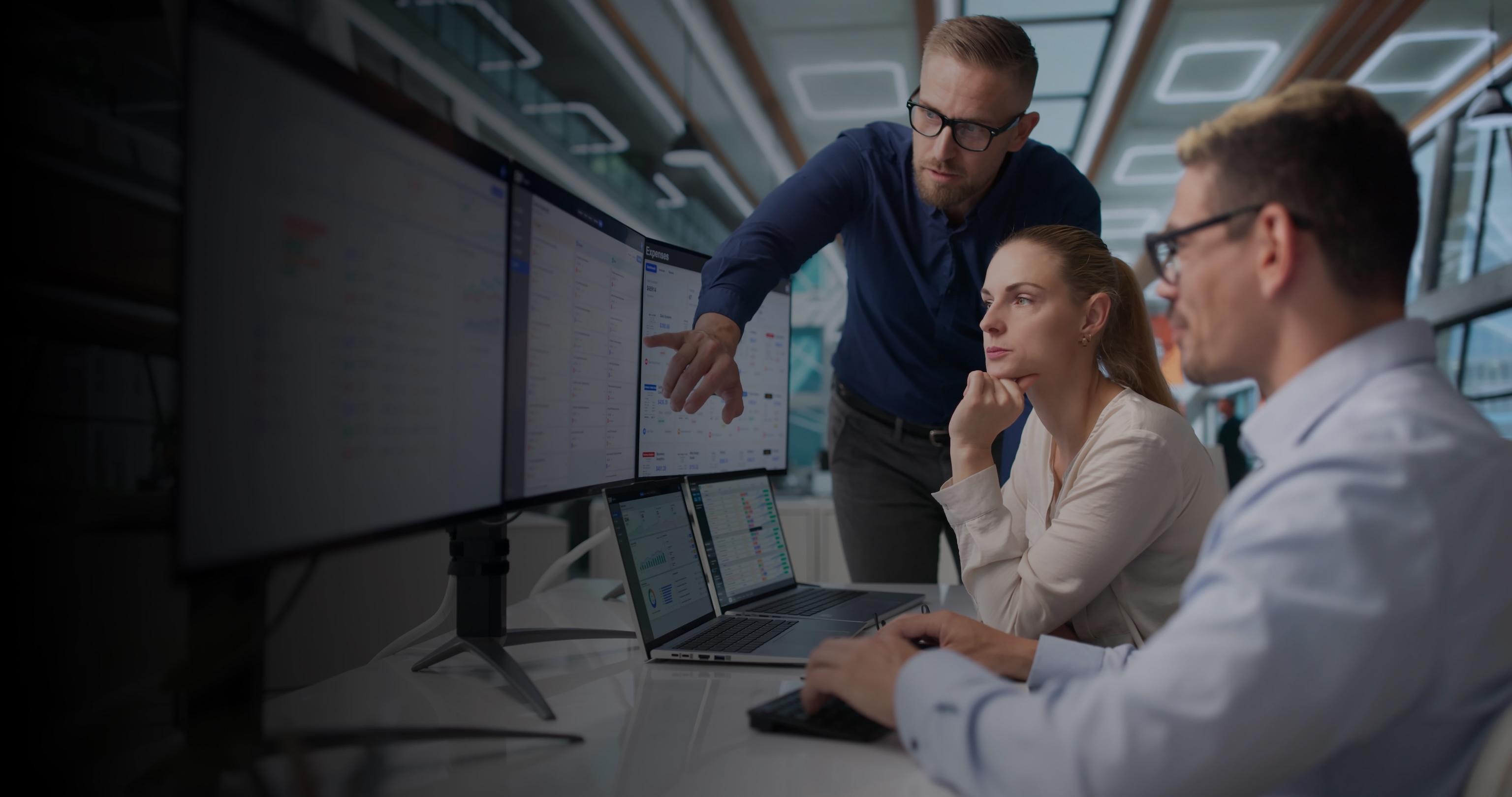 Three professionals collaborate at computer monitors in a modern office, focused on reviewing information in a serious, analytical mood.
