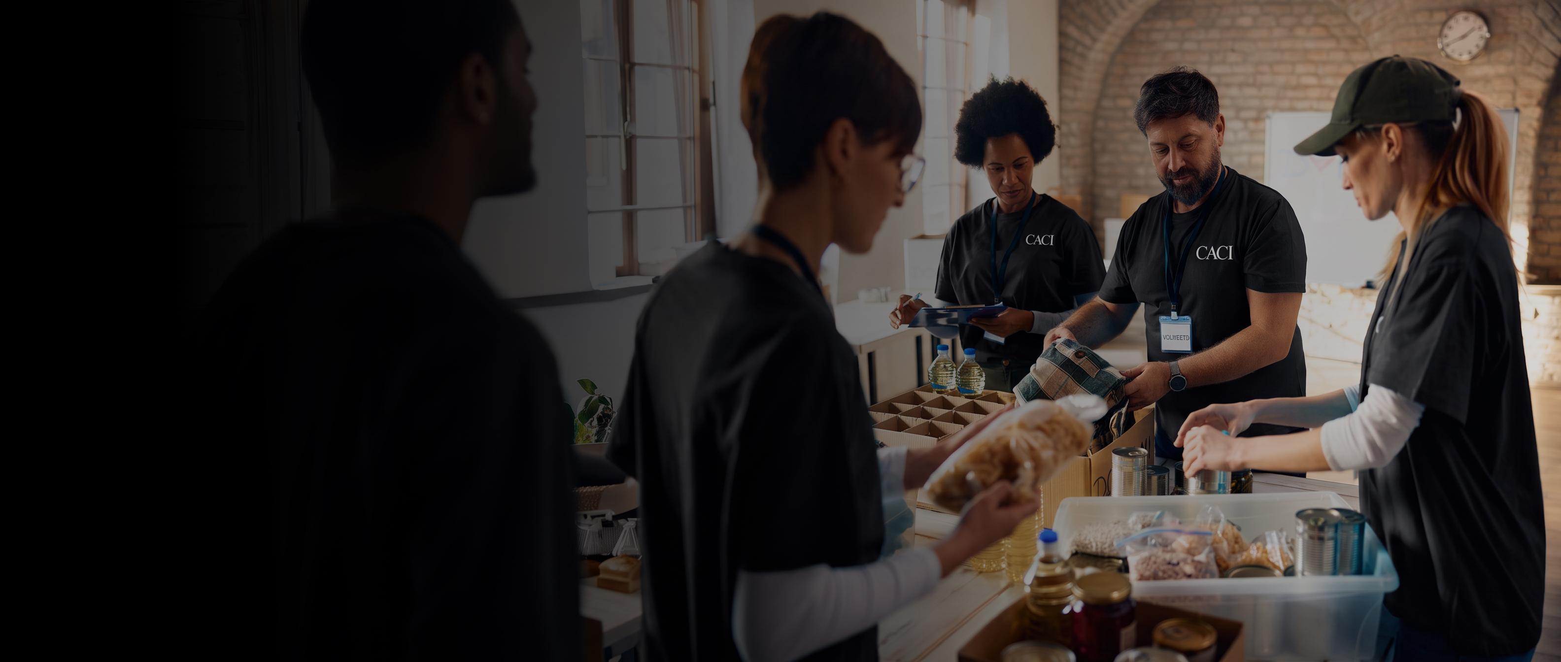 Volunteers prepare and serve food at a community table, working together in a warm kitchen with trays, bread, and drinks.