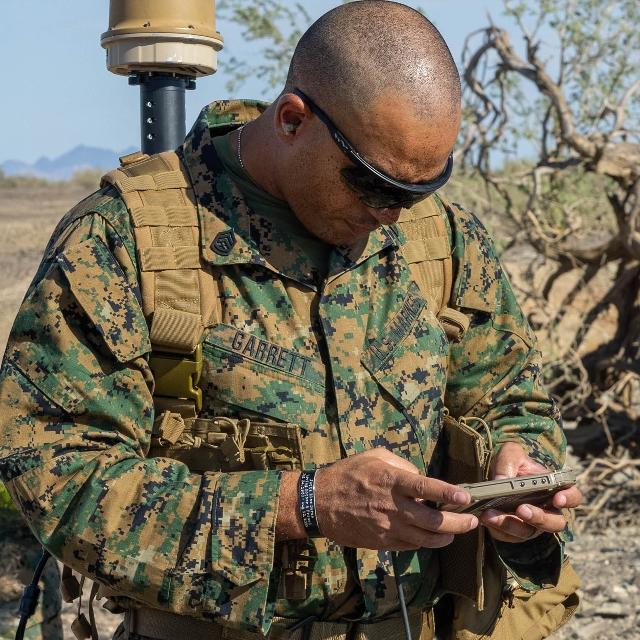 A man in camouflage stands attentively, gazing at his phone in a focused manner.