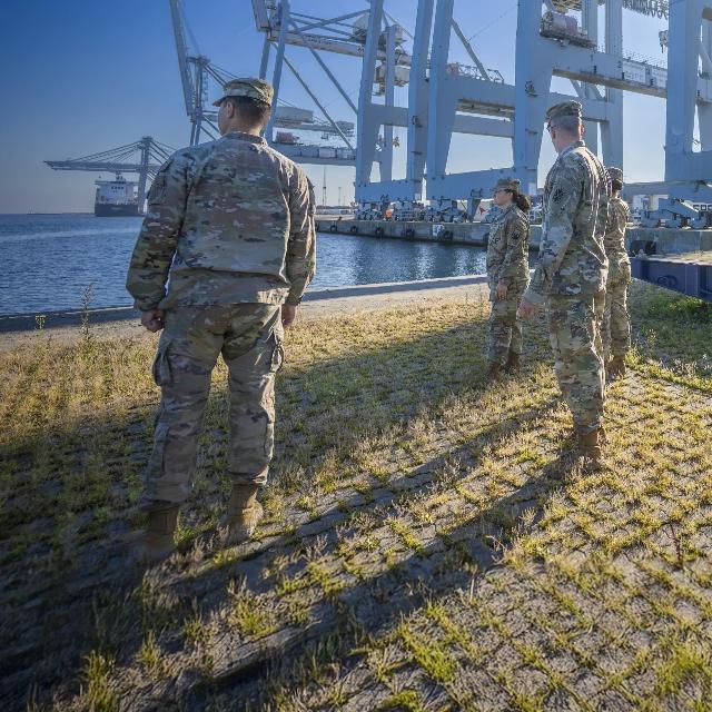 Army soldiers stand on grass beside a large crane, showcasing military presence and equipment in an outdoor setting.