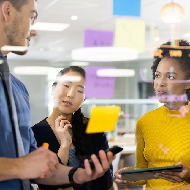 Three people collaborate around a table, with colorful sticky notes displayed on the wall behind them.