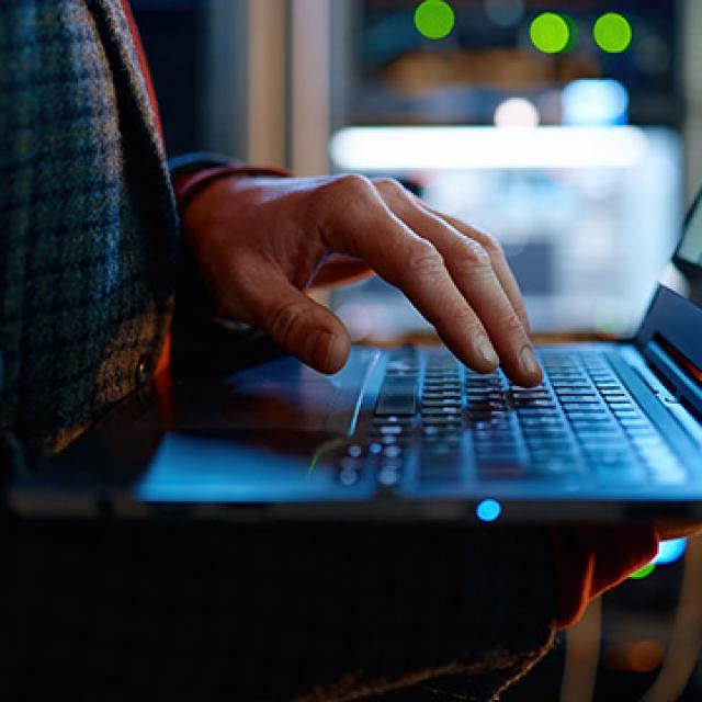 A person in a dark sweater types on a laptop in a dimly lit server room. 
