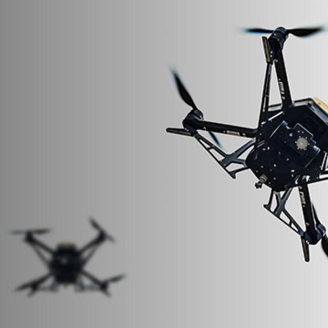 A close-up of a black drone with four spinning propellers in flight against a gradient background.