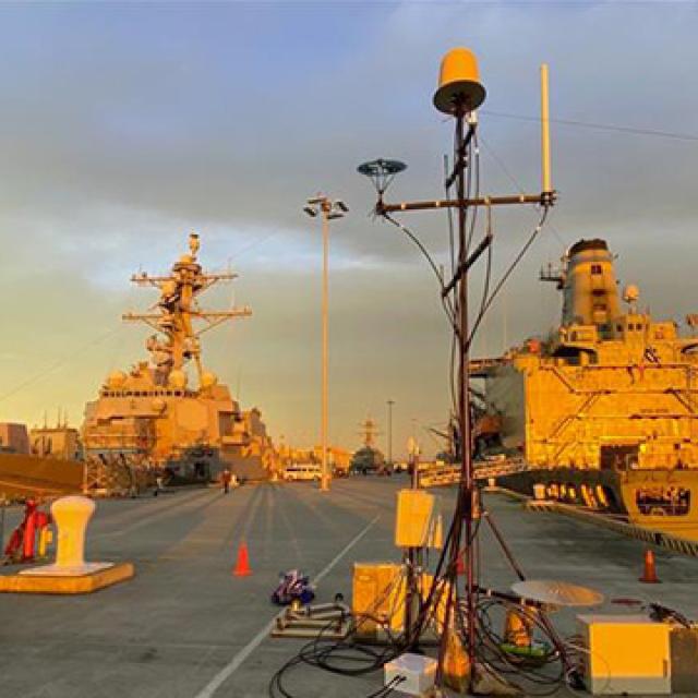 Military ships docked at a harbor during a golden sunset, casting warm light on the scene.