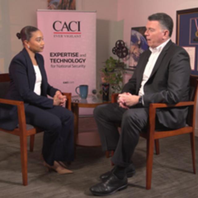 Two professionals seated and talking in an office with awards and a banner displaying "CACI" and "Expertise and Technology for National Security."
