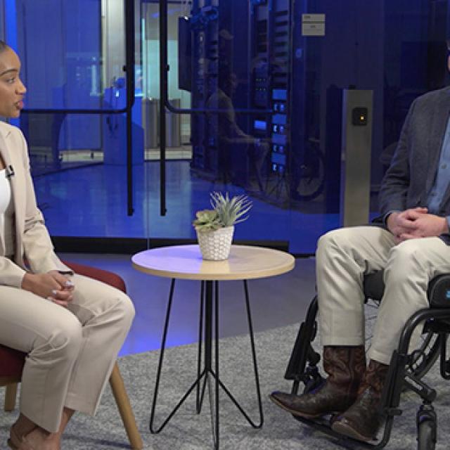 A woman in a beige suit interviews a man in a wheelchair, wearing a blazer and jeans, in a modern office.