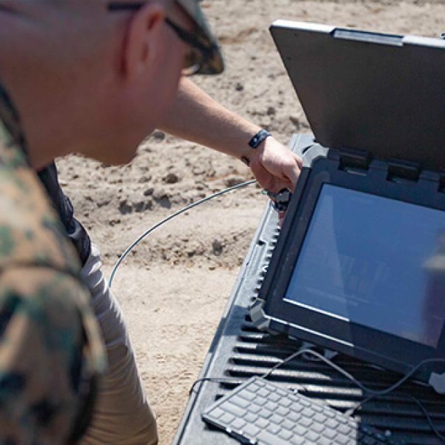 A person in a camouflage uniform operates a rugged laptop on a truck bed in a sandy environment, highlighting outdoor military technology use.