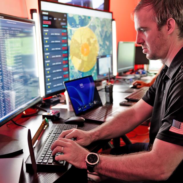 A man seated at a desk, working on two computer monitors with a keyboard in front of him.