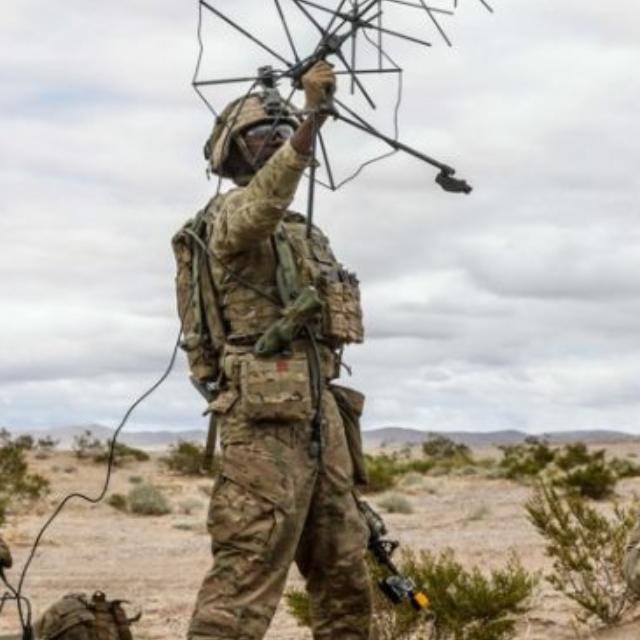 A soldier stands in the desert, holding an antenna, with a clear blue sky in the background.