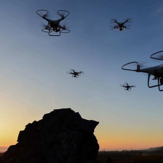 Four drones hover in the evening sky over a silhouetted landscape with a rocky foreground and distant mountains, set against a vibrant sunset.