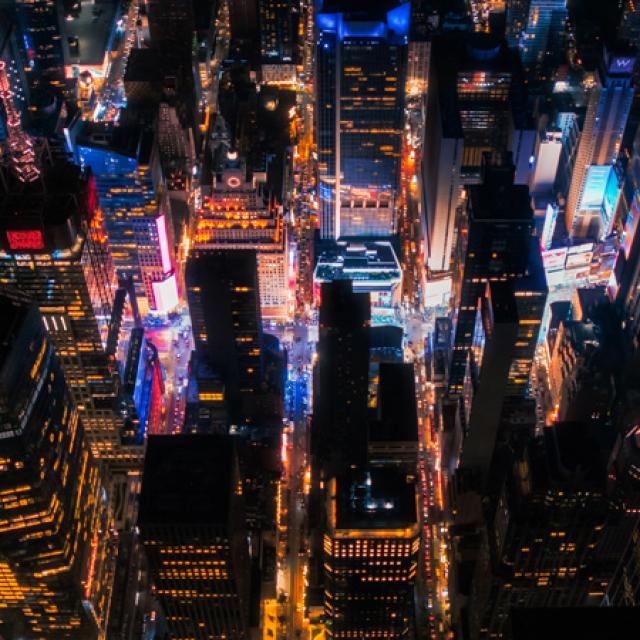 Aerial view of a city at night with vibrant lights illuminating tall skyscrapers in various colors, creating a lively and bustling urban atmosphere.