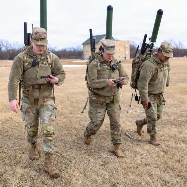 Three soldiers in military uniforms walking through grassy terrain, each carrying their weapons.