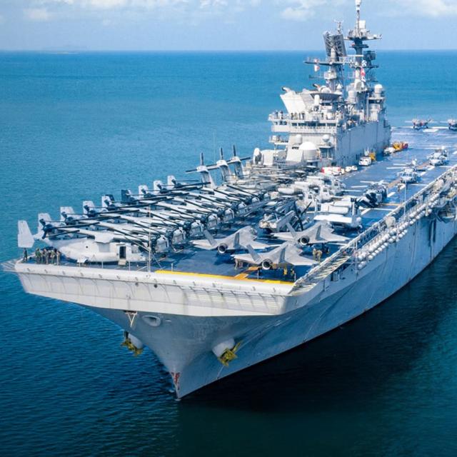 An aircraft carrier sails through the ocean, with waves gently lapping against its hull under a clear blue sky.