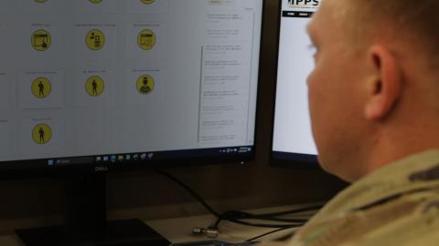 A person in military uniform views a computer screen displaying a grid of yellow icons with various symbols.