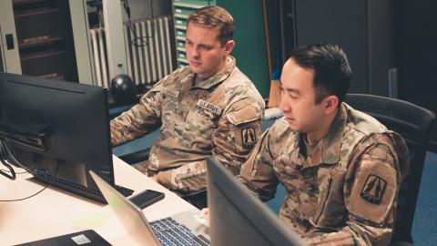Two soldiers in uniform seated at computers, focused on their tasks in a military setting.