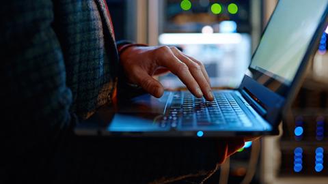 A person in a dark sweater types on a laptop in a dimly lit server room. 