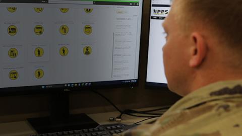 A person in military uniform views a computer screen displaying a grid of yellow icons with various symbols.