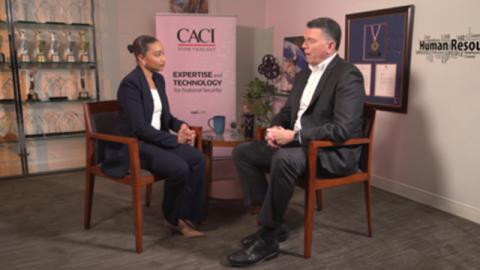 Two professionals seated and talking in an office with awards and a banner displaying "CACI" and "Expertise and Technology for National Security."rity.