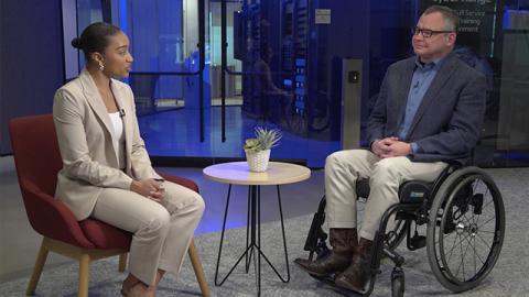A woman in a beige suit interviews a man in a wheelchair, wearing a blazer and jeans, in a modern office.