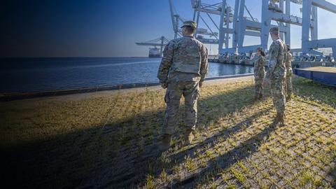 Four soldiers in camouflage uniforms stand on a grassy slope overlooking a shipping port.
