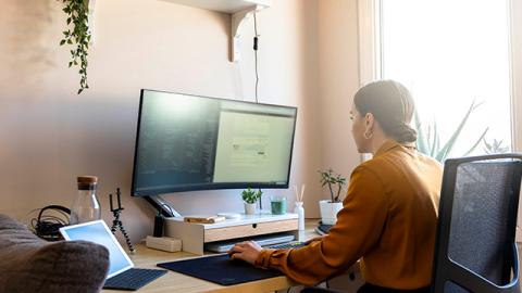 A woman in a mustard blouse works at a desk with a large monitor displaying code and documents.