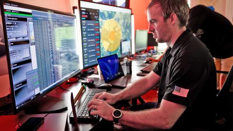A man seated at a desk, working on two computer monitors with a keyboard in front of him.