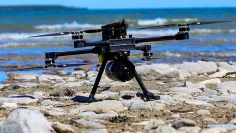 A small drone rests on the sandy beach, with gentle waves lapping in the background.