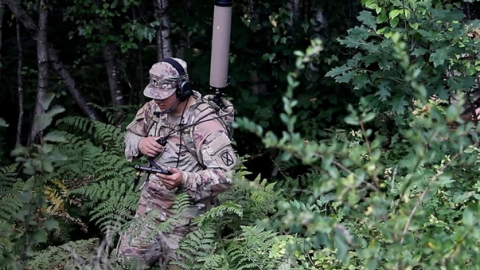 A soldier in camouflage walks through a dense forest, blending into the natural surroundings.
