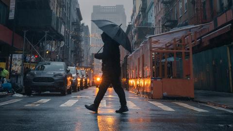 A person walks down a city street, holding an umbrella against the rain, surrounded by buildings and pedestrians.