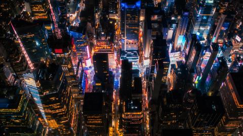 Aerial view of a city at night with vibrant lights illuminating tall skyscrapers in various colors, creating a lively and bustling urban atmosphere.
