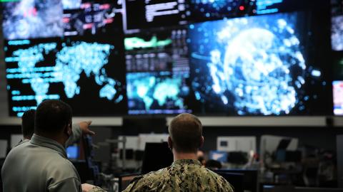 Two military personnel confer in a command center before glowing screens showing world maps, data feeds, and a digital globe; serious tone.