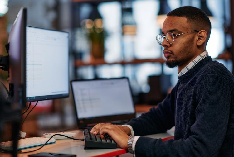 A skilled programmer sits at his computer in a modern office, immersed in problem-solving and showcasing his expertise in technology.