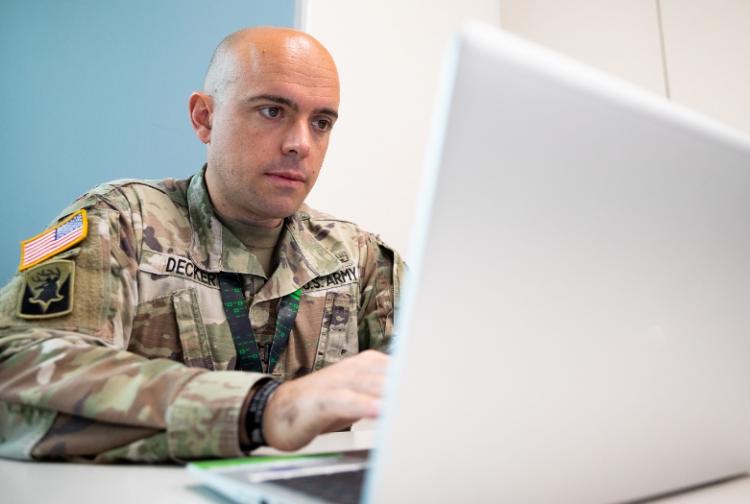 A soldier in uniform is focused on using a laptop, showcasing technology in a military setting.