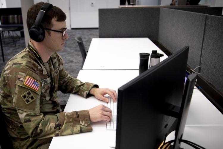 A soldier in uniform sits at a desk, wearing headphones, focused on a task in a professional setting.