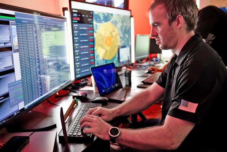 A man seated at a desk, working on two monitors with a keyboard in front of him.