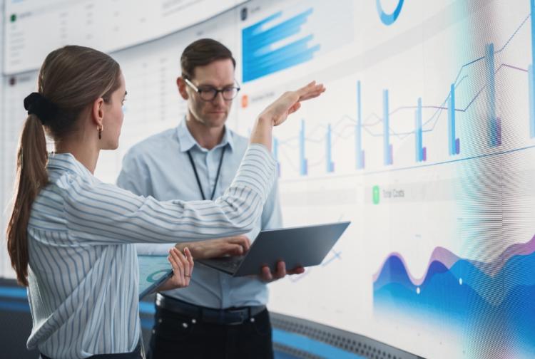 Two individuals analyzing data displayed on a large screen filled with various graphs and charts.