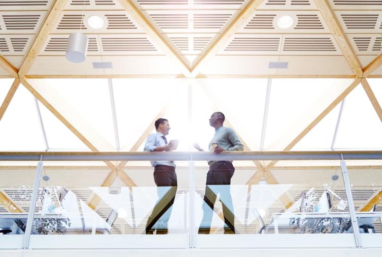 Two individuals standing in a modern open office space, engaged in conversation near a large window.
