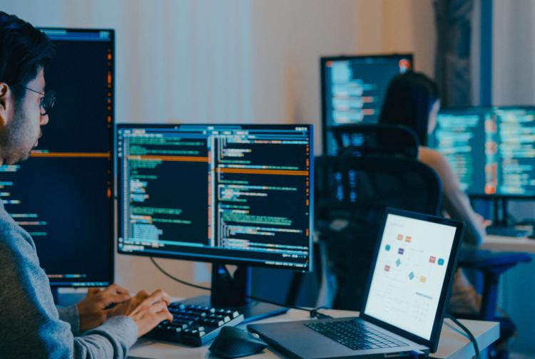 Two software developers work at desks with code displayed across multiple monitors in a modern office, creating a focused, collaborative atmosphere.