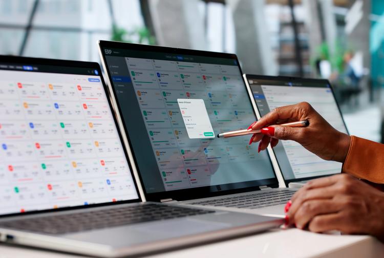 Three laptops display colorful analytics dashboards as a professional points with a red pen; a bright office conveys digital productivity.